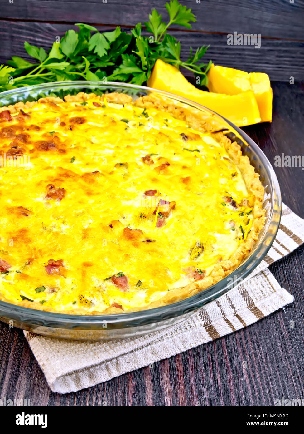 Pumpkin kish with pumpkin and bacon, filled with milk and eggs in a glass frying pan on a napkin, parsley, vegetable slices against a dark wooden boar Stock Photo