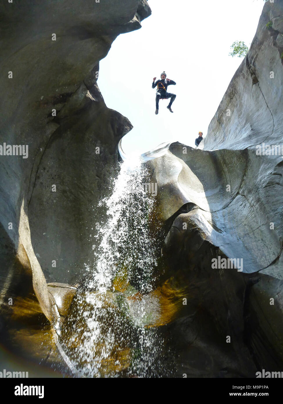 A man in wetsuit jumping from a high cliff next to a waterfall during a canyoning adventure trip in Switzerland. Ticino, in the southern part of Switzerland is a small paradise for canyoning thanks to numerous little waterfalls, torrents and rivers that rush down the valley through the wilderness. Cresciano, Ticino Canton, Switzerland Stock Photo