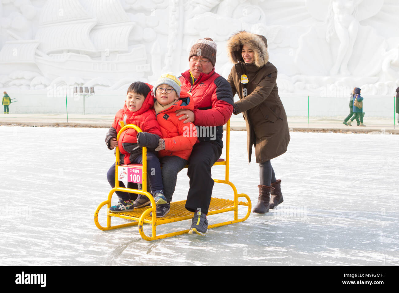 An Asian family is having fun on a group sled at a frozen river.  The Hwacheon Sancheoneo Ice Festival is a tradition for Korean people. Every year in January crowds gather at the frozen river to celebrate the cold and snow of winter. Main attraction is ice fishing. Young and old wait patiently over a small hole in the ice for a trout to bite. In tents they can let the fish grilled after which they are eaten. Among other activities are sledding and ice skating.  The nearby Pyeongchang region will host the Winter Olympics in February 2018. Stock Photo