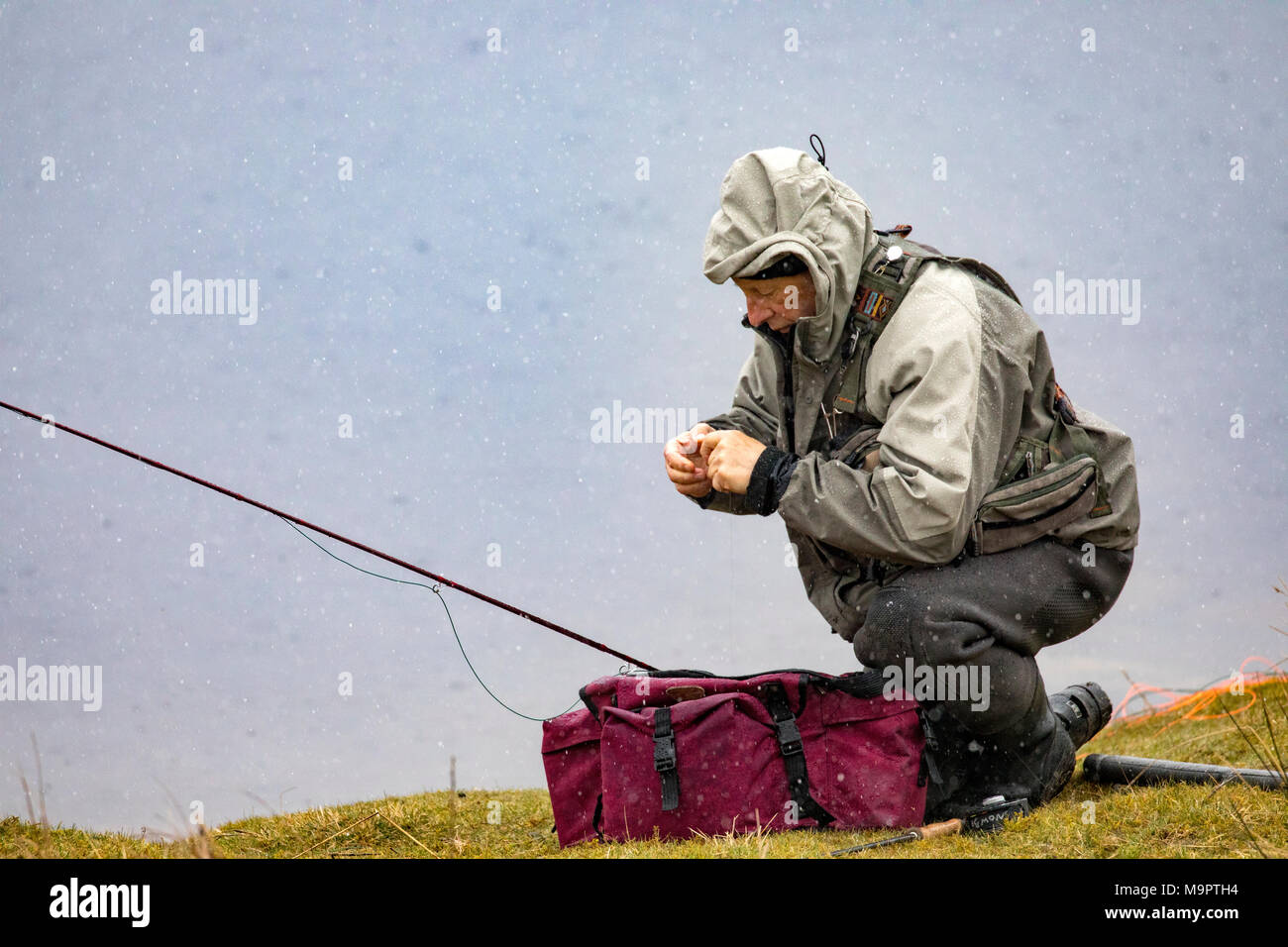 North Wales, UK 28th March 2018, UK Weather:  A further spell of snowfall and colder temperatures hits North Wales with further snow expected in the coming week. A fly fisherman tying on a new fly as the snow begins to fall on Llyn Brenig, Conwy County, North Wales © DGDImages/Alamy Live News Stock Photo