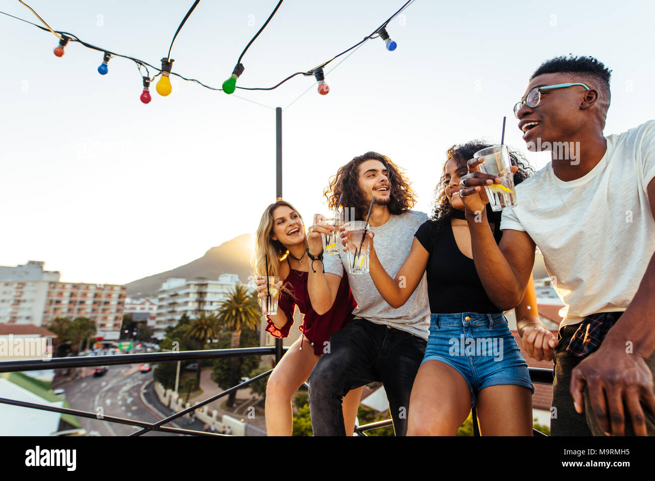 Four young people drinking and smiling on the rooftop in evening. Men and women partying with drinks on terrace. Stock Photo