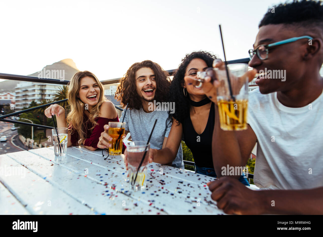 Multi-ethnic group of friends sitting on the rooftop with drinks. Men and women partying on the rooftop in evening. Stock Photo