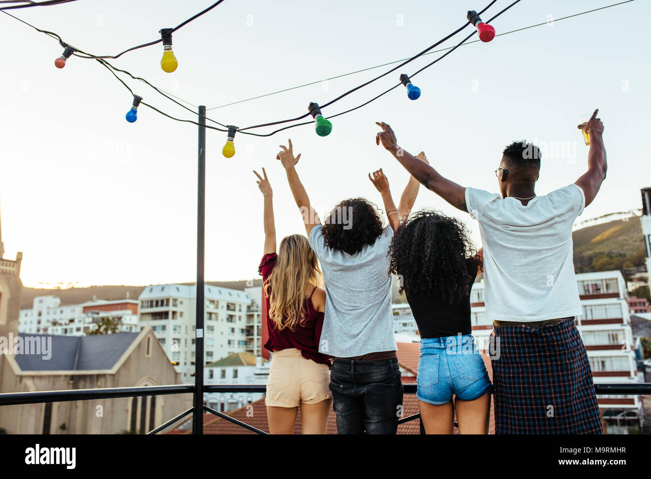 Men and woman partying on terrace with drinks at sunset. Rear view of friends standing with hands raised on rooftop. Stock Photo