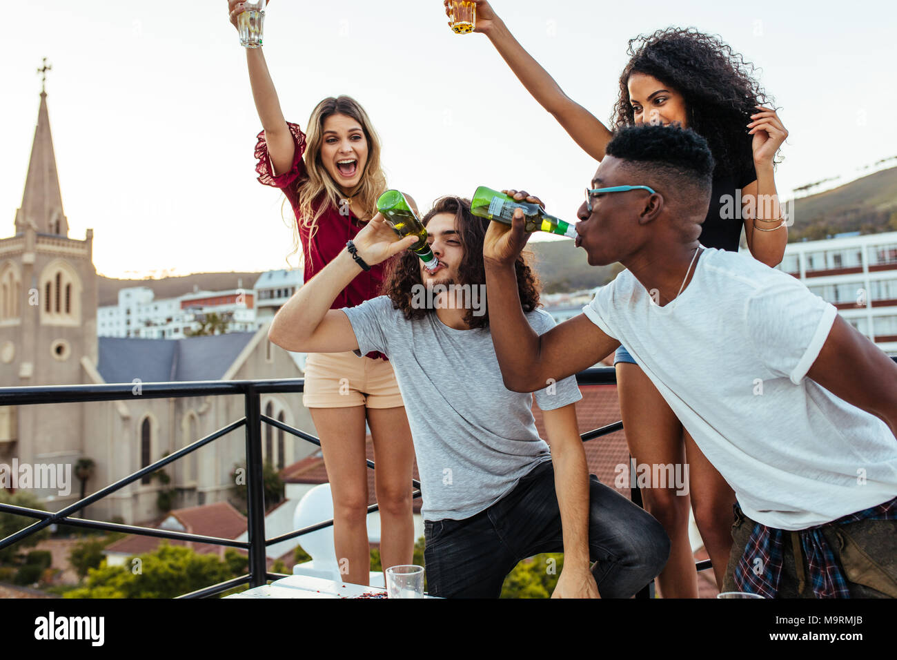 Multiracial young people partying on terrace, with men drinking beers together and girls cheering. Beer drinking challenge at rooftop party. Stock Photo