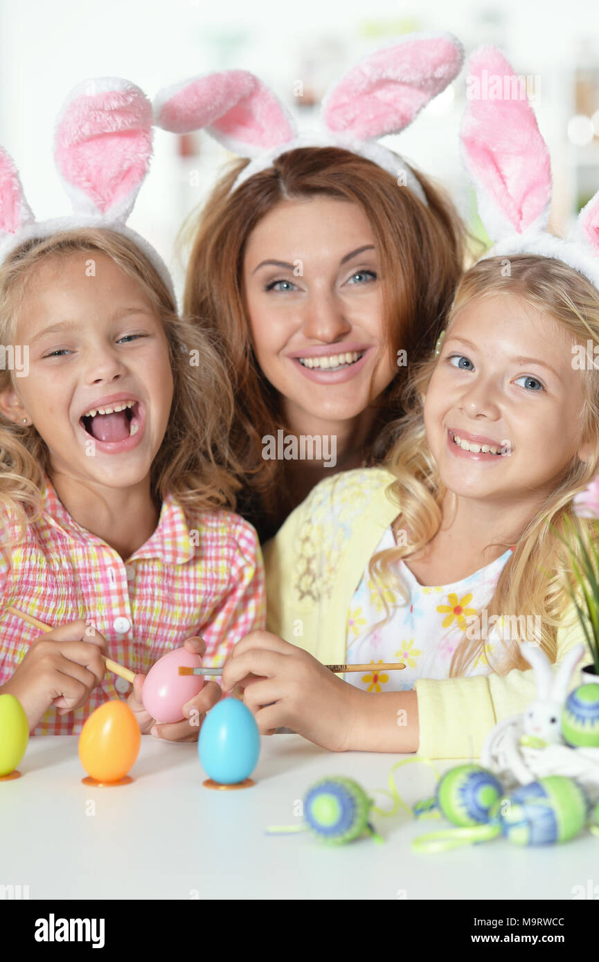 Happy young mother with her cute daughters wearing rabbit ears and painting Easter eggs at home Stock Photo