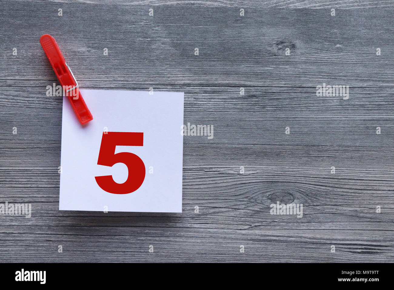 Series of red numbers on white note paper, held by red clothes peg, on grey plank background with space for copy Stock Photo