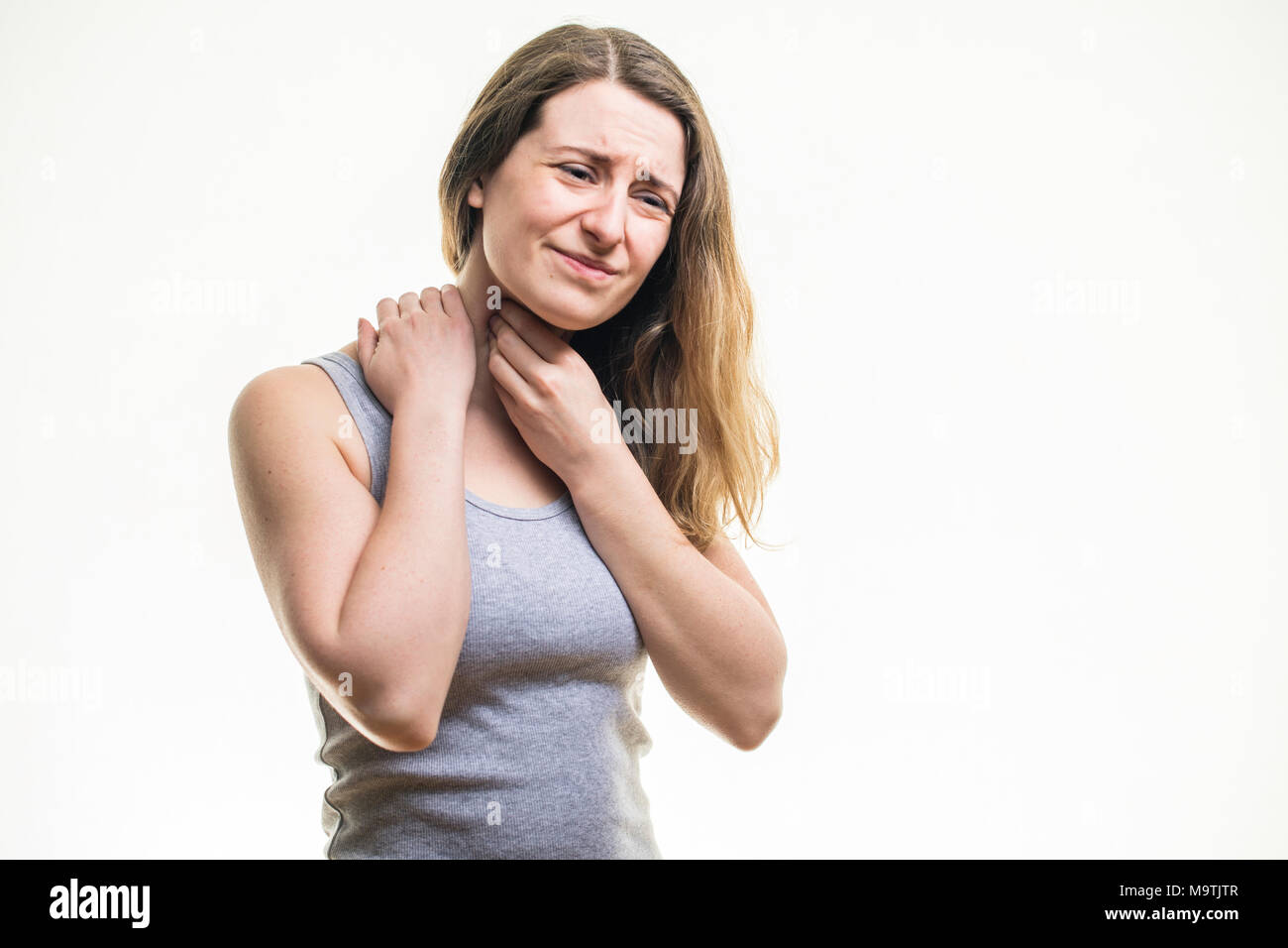 A young Caucasian woman girl wearing a grey vest,  suffering from a sore throat or neck pain, rubbing her skin for relief,  standing against a white background, UK Stock Photo