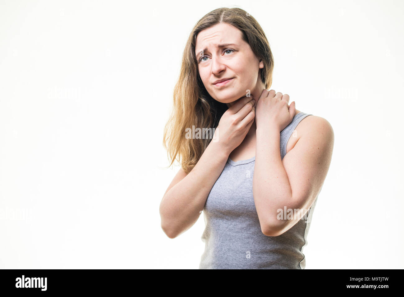 A young Caucasian woman girl wearing a grey vest,  suffering from a sore throat or neck pain, rubbing her skin for relief,  standing against a white background, UK Stock Photo