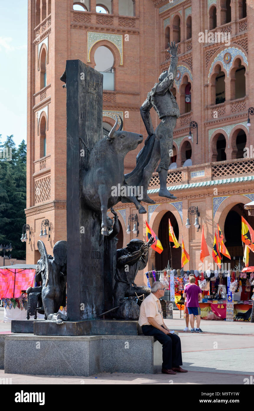 Old man sitting on a bullfighter statue in plaza in front of Madrid's Las Ventas bullring with multiple Spanish flags, stalls and people Stock Photo