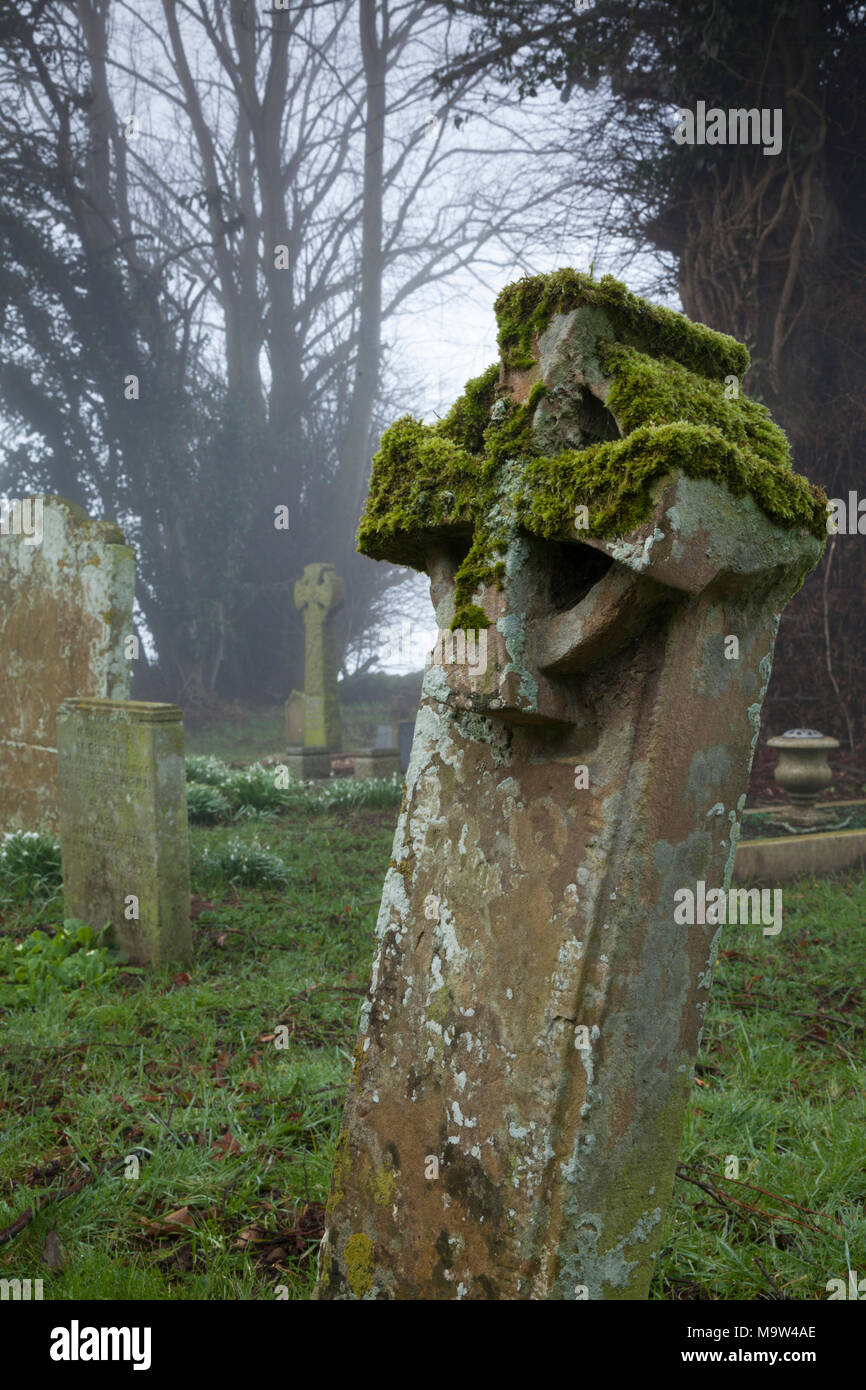 A stone Celtic Cross gravestone with moss and lichen on a misty winter morning, All Saints church, Holdenby, Northamptonshire, England. Stock Photo