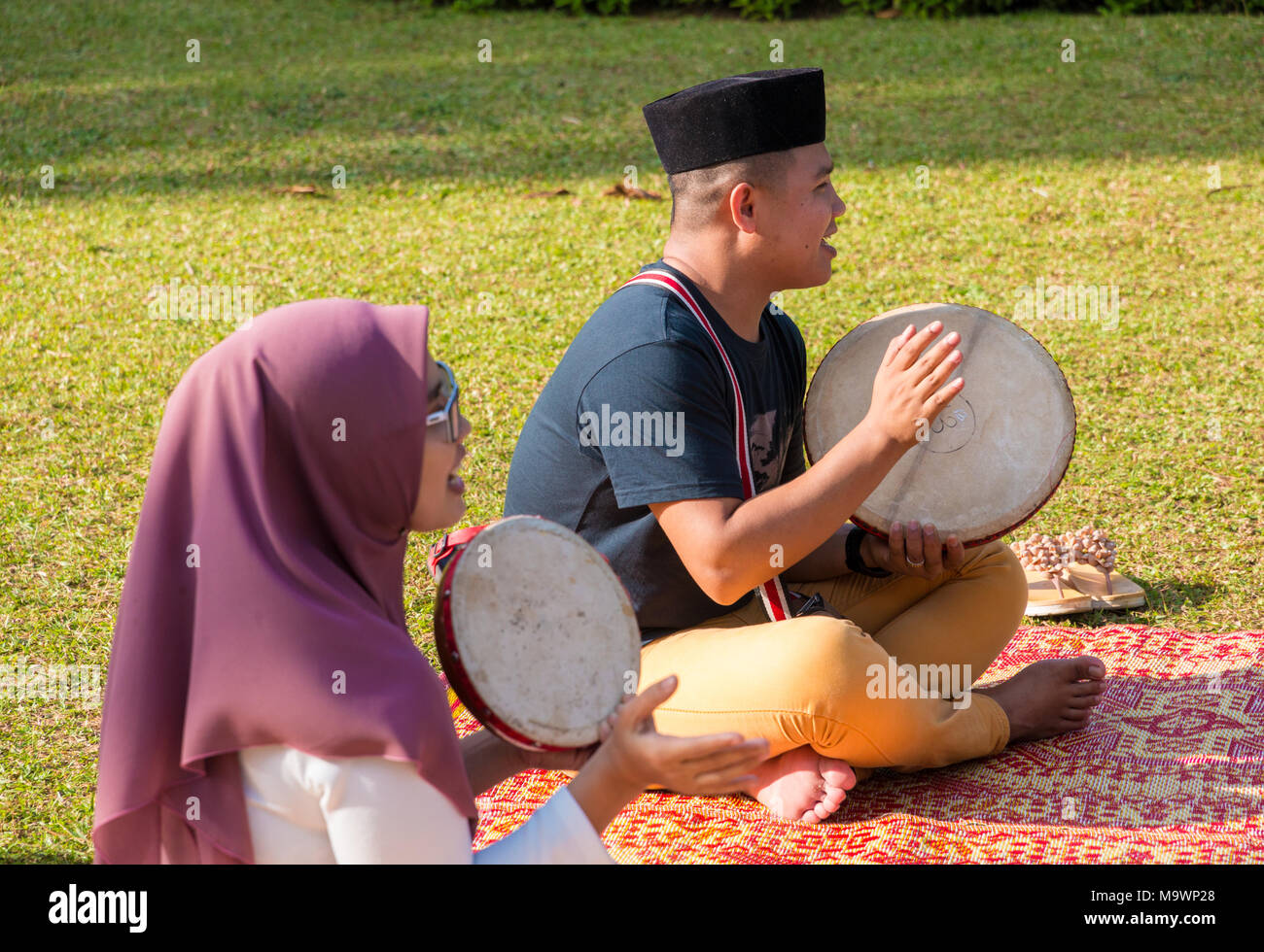 Two Malaysians, a man with a sonkok and a woman wearing a hijab, are sitting on a traditional rattan mat and playing the kompang (Malay drum). Stock Photo