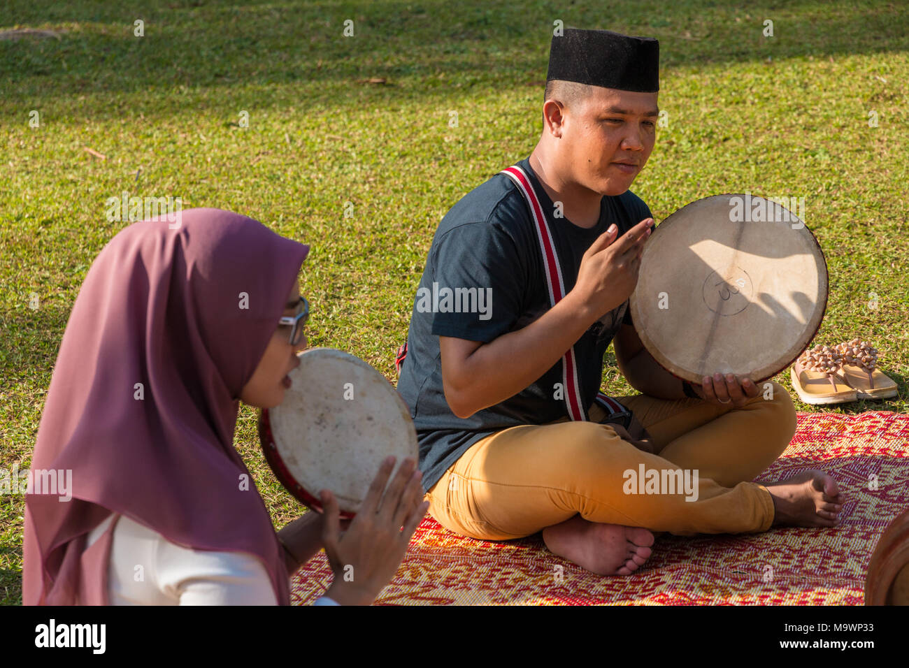 A Malay man with a sonkok and a Malay woman wearing a hijab are sitting on a traditional rattan mat and showing how to play the kompang (Malay drum). Stock Photo