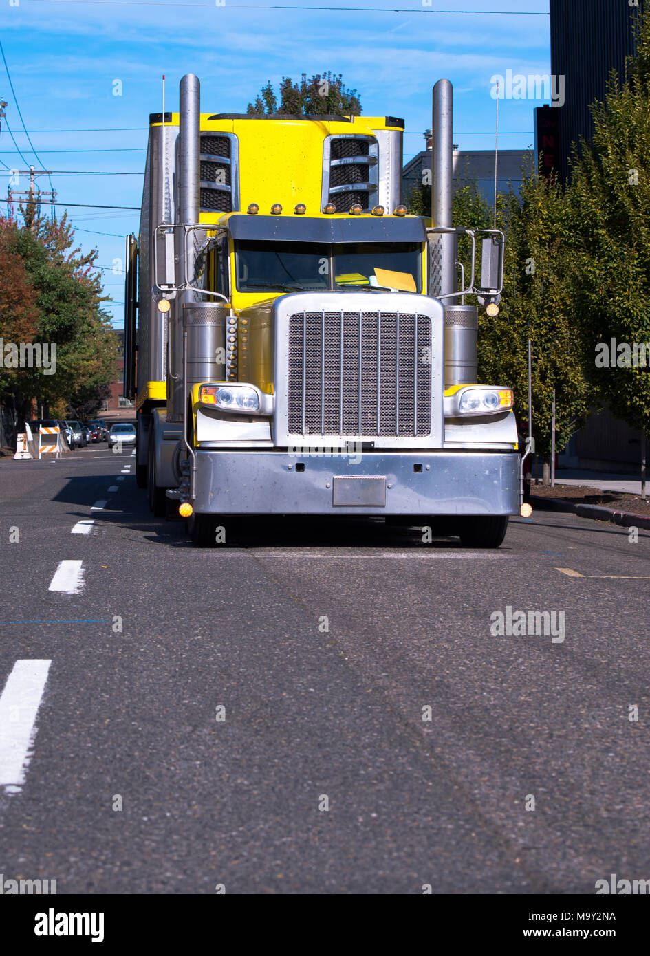 Bright yellow American classic big rig semi truck fleet with freezer semi trailer and tall chrome exhaust pipes running by urban city street for timel Stock Photo