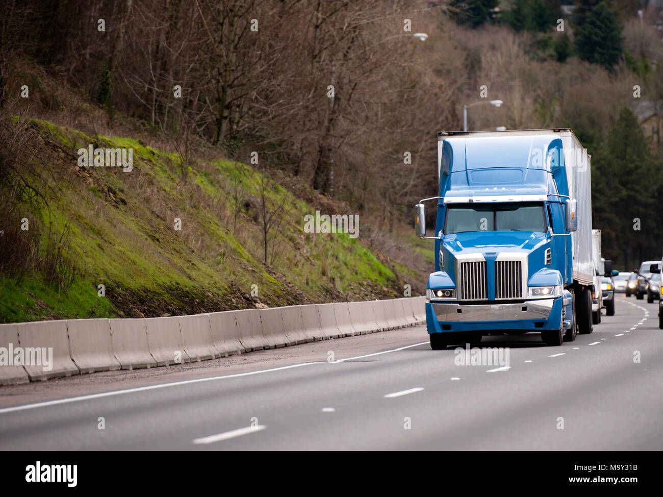 Modern blue powerful stylish big rig tall cab semi truck fleet for long haul deliveries with semi trailer going on wide highway with trees on the hill Stock Photo