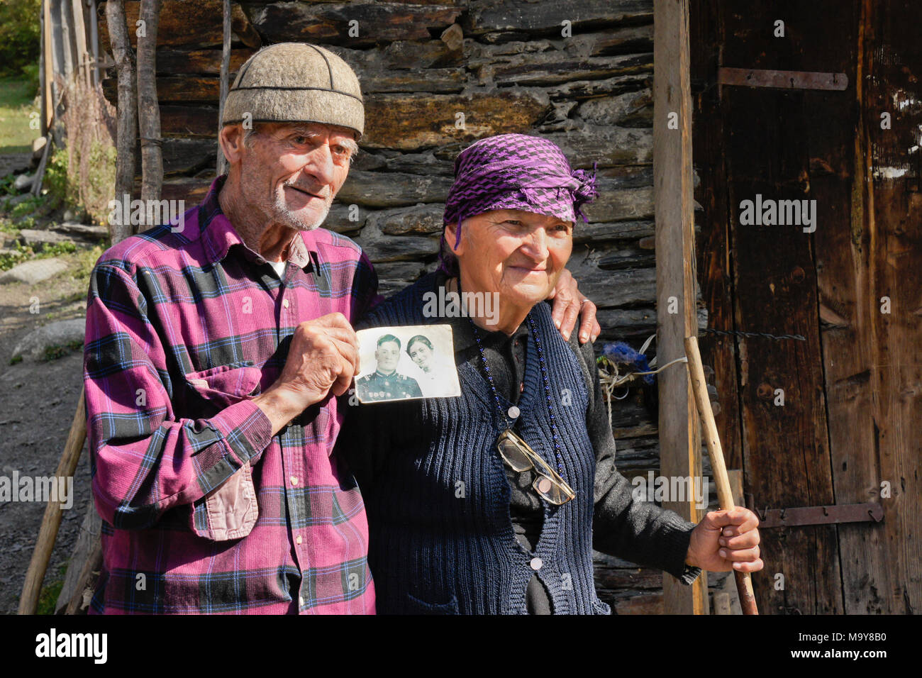 An elderly married couple standing outside an old stone shed display a photograph of themselves, taken in 1958, Ushguli, Upper Svaneti, Georgia Stock Photo