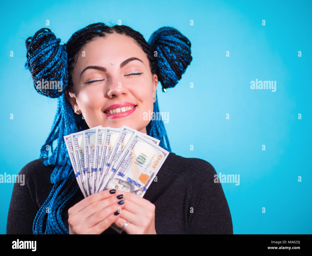 Happy smiling woman holding cash money dollars and thinking how to spent its. Toned portrait. Hipster girl with blue dyed braids african hairstyle in studio. Stock Photo