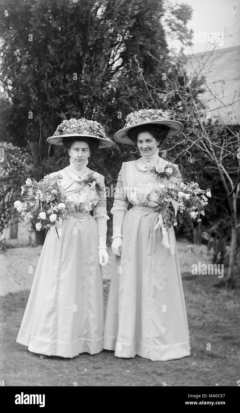 Two women dressed as bridesmaids in late Victorian England. Both wear white dresses with elaborate white flowered hats, and both carry a large posy of flowers. Stock Photo