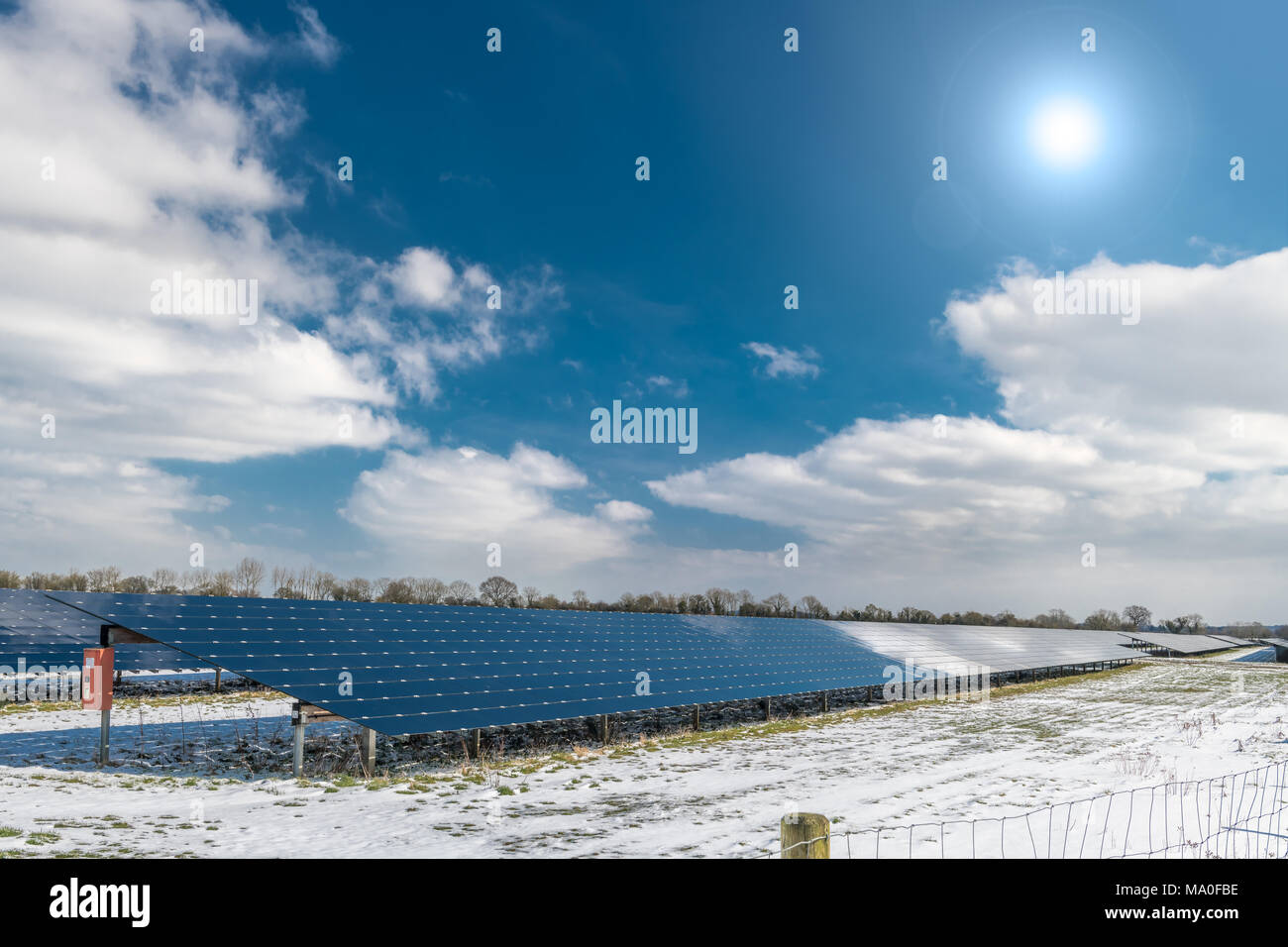 The sun shines above a snow covered field containing an array of Solar Panels. Stock Photo