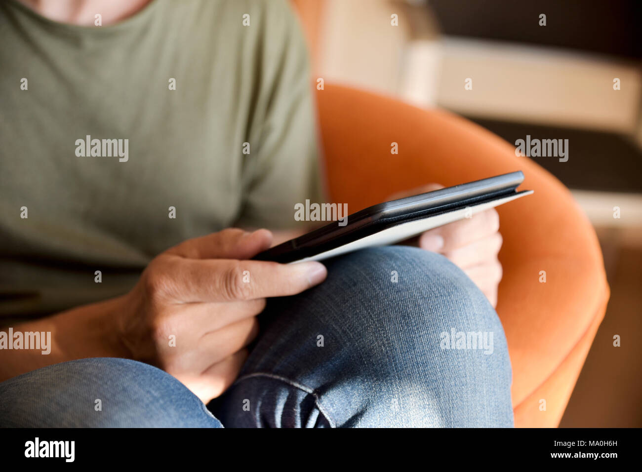 closeup of a young caucasian, wearing jeans and a green t-shirt, using a tablet sitting in a comfortable orange couch indoors Stock Photo