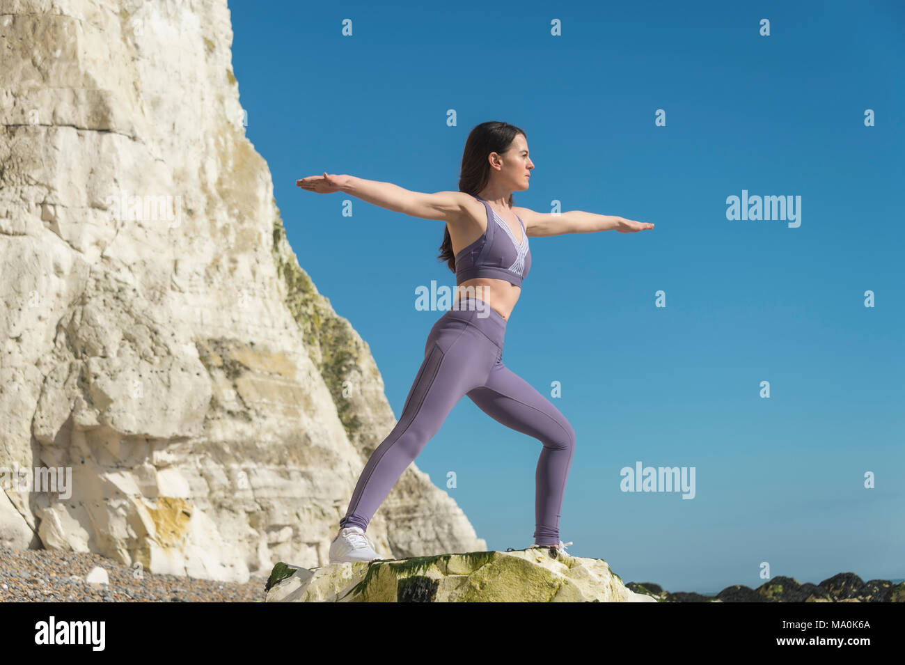 woman in the 'warrior II' yoga pose, standing on rocks by the sea with