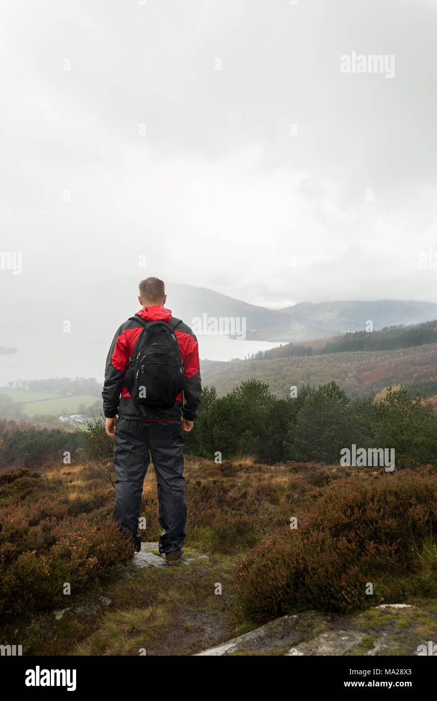 Man dressed for wet weather hiking stands on top of a hill looking across a cloudy autumn hilly landscape scene of Loch Lomand National Park, Scotland Stock Photo