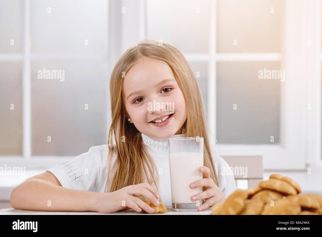 Smiling little girl is drinking milk from transparent glass with delicious  cookies,which lay on brown patterned plate. Child wears white sweatshirt  and has long blonde hair. Window on background Stock Photo -
