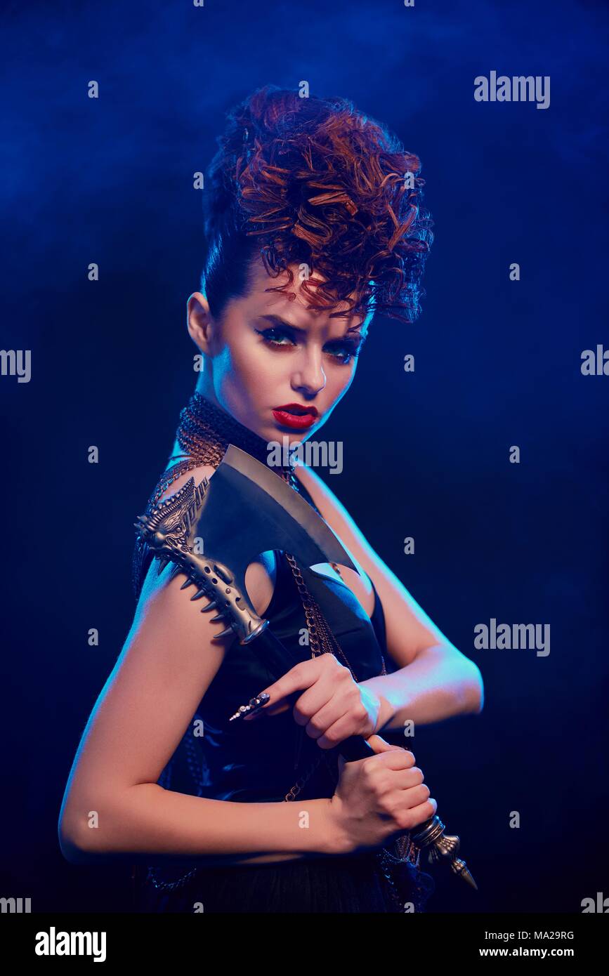 Side view of fantastic girl keeping sharp metallic axe with thorns. Female warrior standing on dark blue smoky background. Model wearing black top with opened shoulders, make up and stylish hairdress. Stock Photo