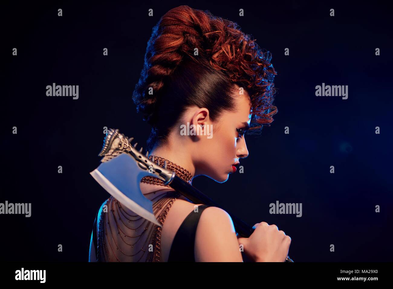 Back view of young beatiful warrior girl keeping sharp metallic axe with thorns. Model wears bright original make up and fashionable, stylish hairdress. Golden chains with crosses on neck. Stock Photo