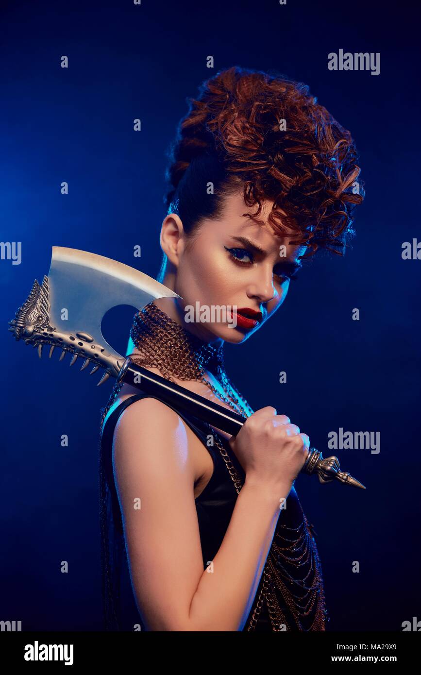 Close up of dangerous beatiful girl keeping sharp metallic axe with thorns. Model wears black top with opened shoulders,bright make up and stylish hairdress. She stands on dark blue smoky background. Stock Photo