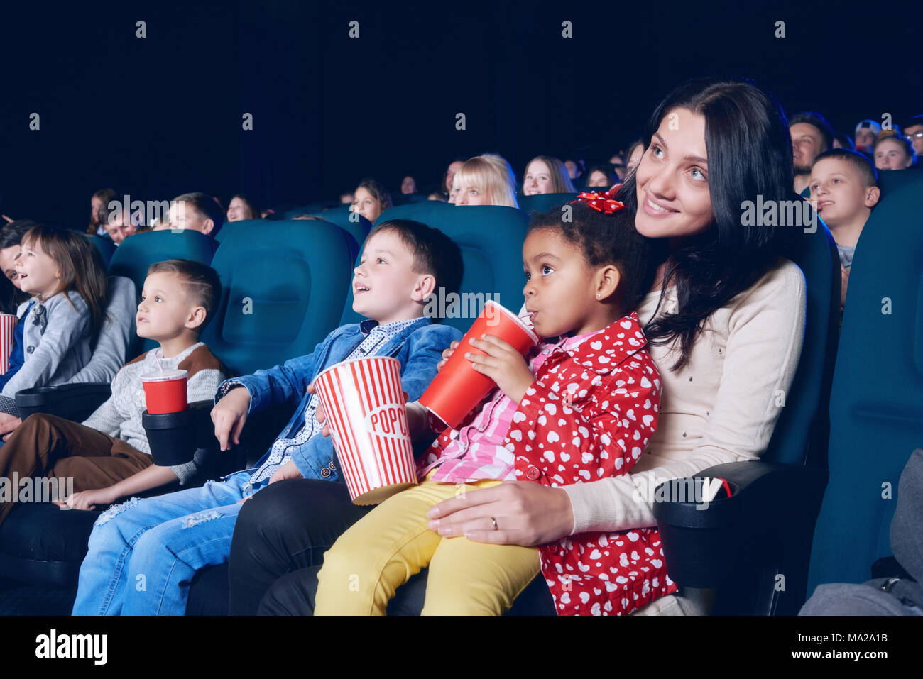 Sideview of nice mother and daughter watching interesting movie in cinema hall on other people's background. Little girl wearing colorful clothes, drinking cola and eating popcorn. Stock Photo