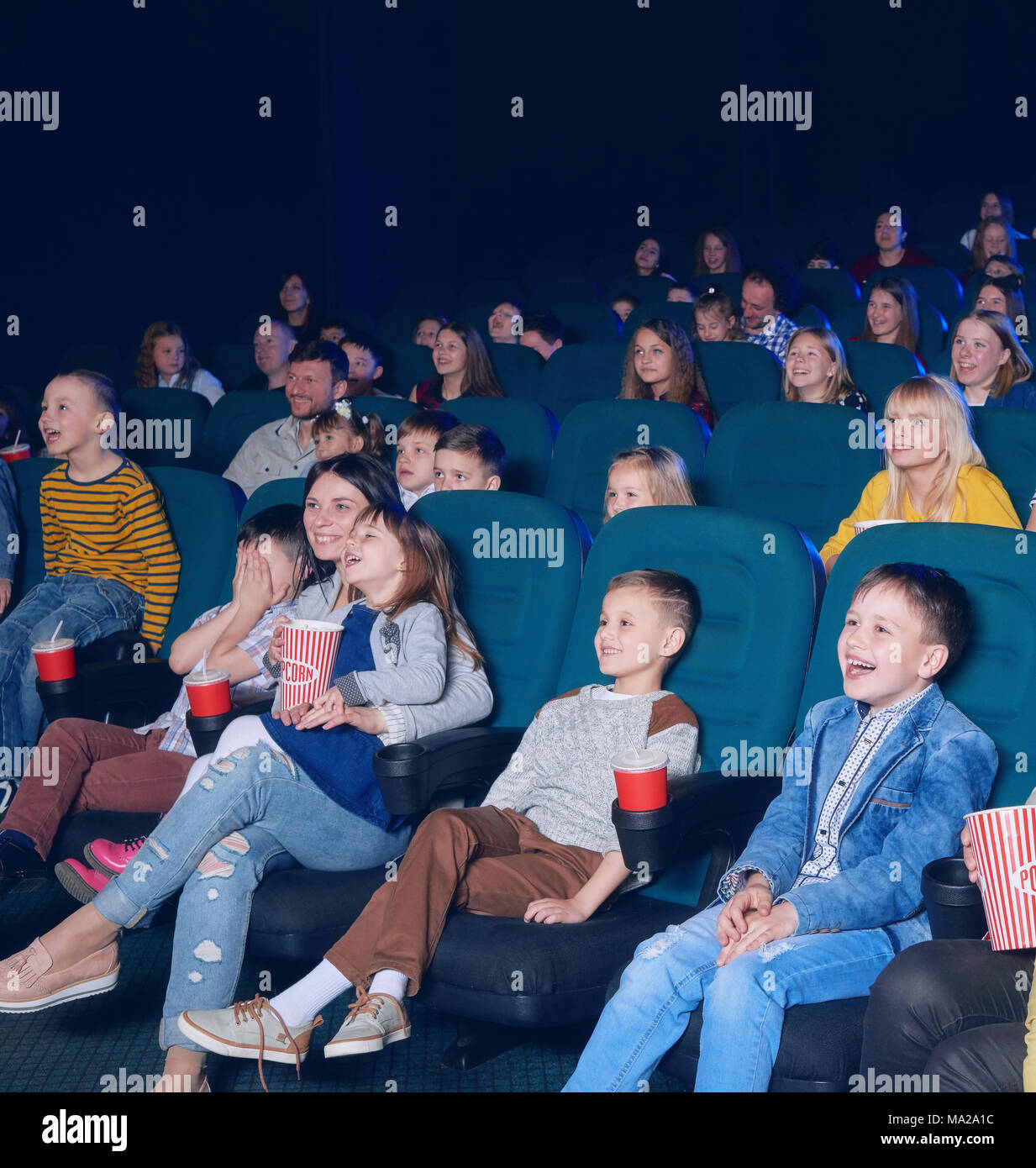 sideview of happy children watching interesting, funny movie, sitting on comfortable green places. Boys and girls laughing, smiling, eating popcorn and drinking fizzy drinks. Stock Photo