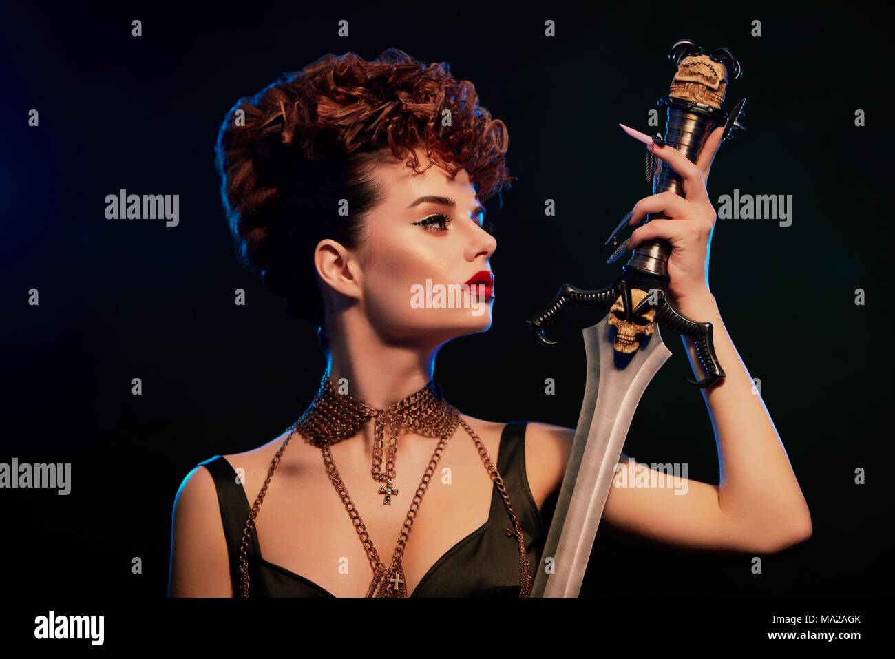 Fashionable young model with bright make up keeping steel sword with black skull. Woman wearing stylish hairdress with little curles. Female in black top with thin chains. Confident original. Stock Photo