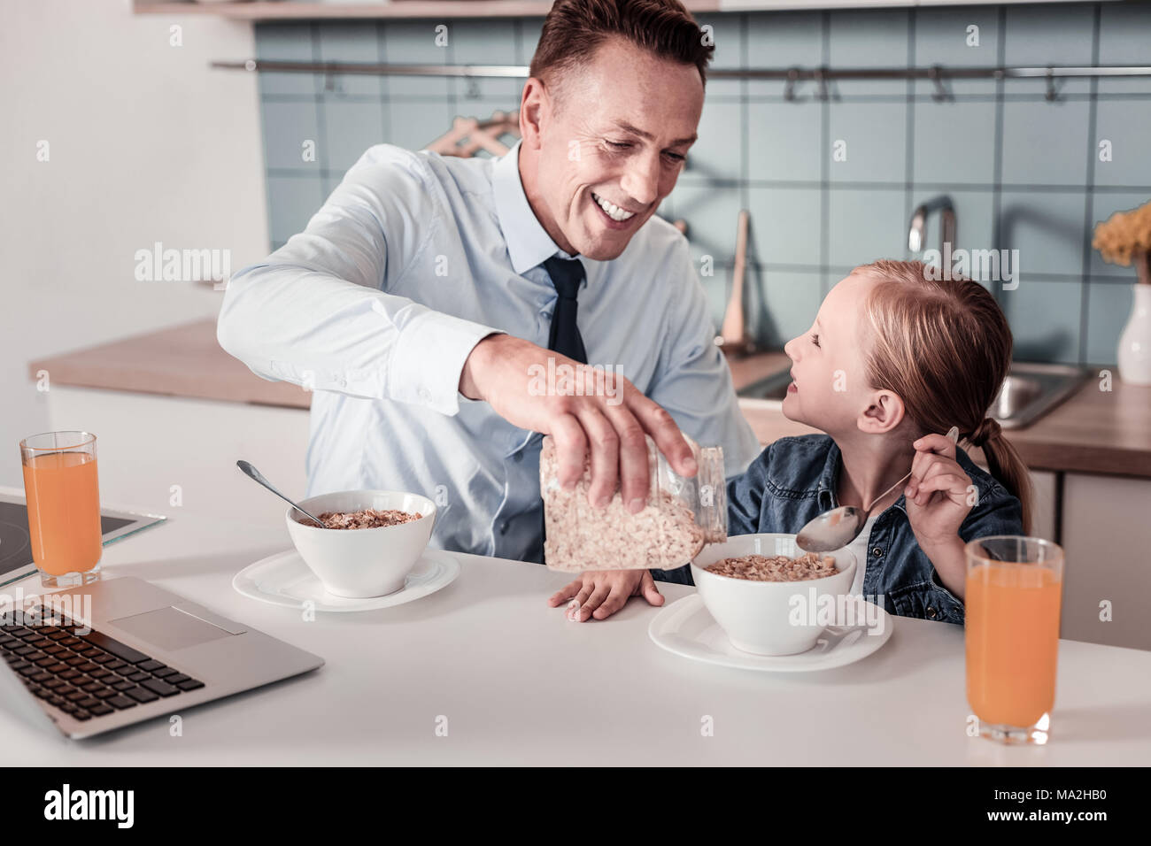 Healthy food. Cheerful man keeping smile on his face and pouring down corn flakes while talking with his daughter Stock Photo