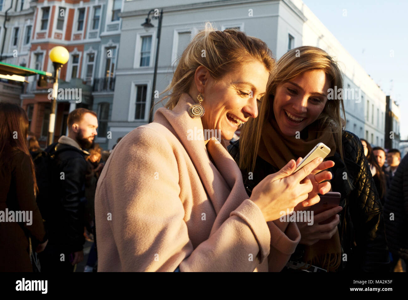 Young female adults smiling with emotion, looking at mobile phone smart phone. Communication. Millennials expressing emotion. People happy street. Stock Photo