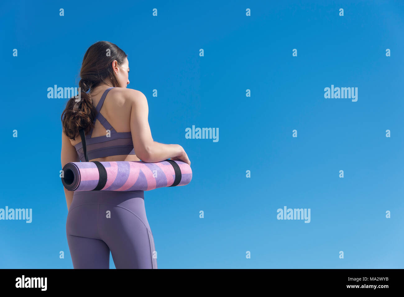 back view of a woman carrying a rolled up yoga mat with blue sky background Stock Photo