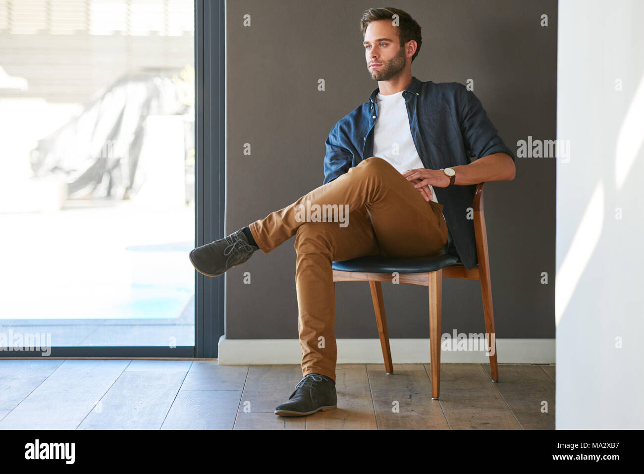 Fashionable young professional looking off into the distance while seated in a beautiful wooden chair in his modern home with a lovely grey textured w Stock Photo