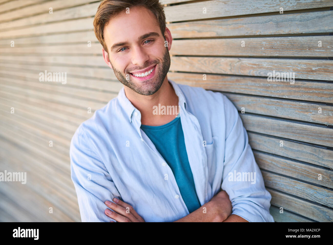Tightly cropped image of handsome young male in his mid 20s smiling at camera with his teeth showing and his arms crossed while leaning against beauti Stock Photo