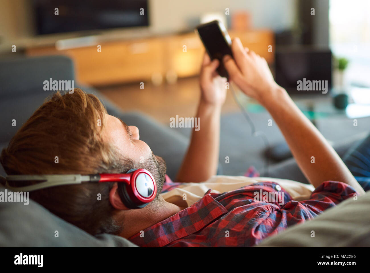 Over the shoulder image of a young caucasian guy listening to music through headphones from his phone that he is holding in his hands, all while relax Stock Photo