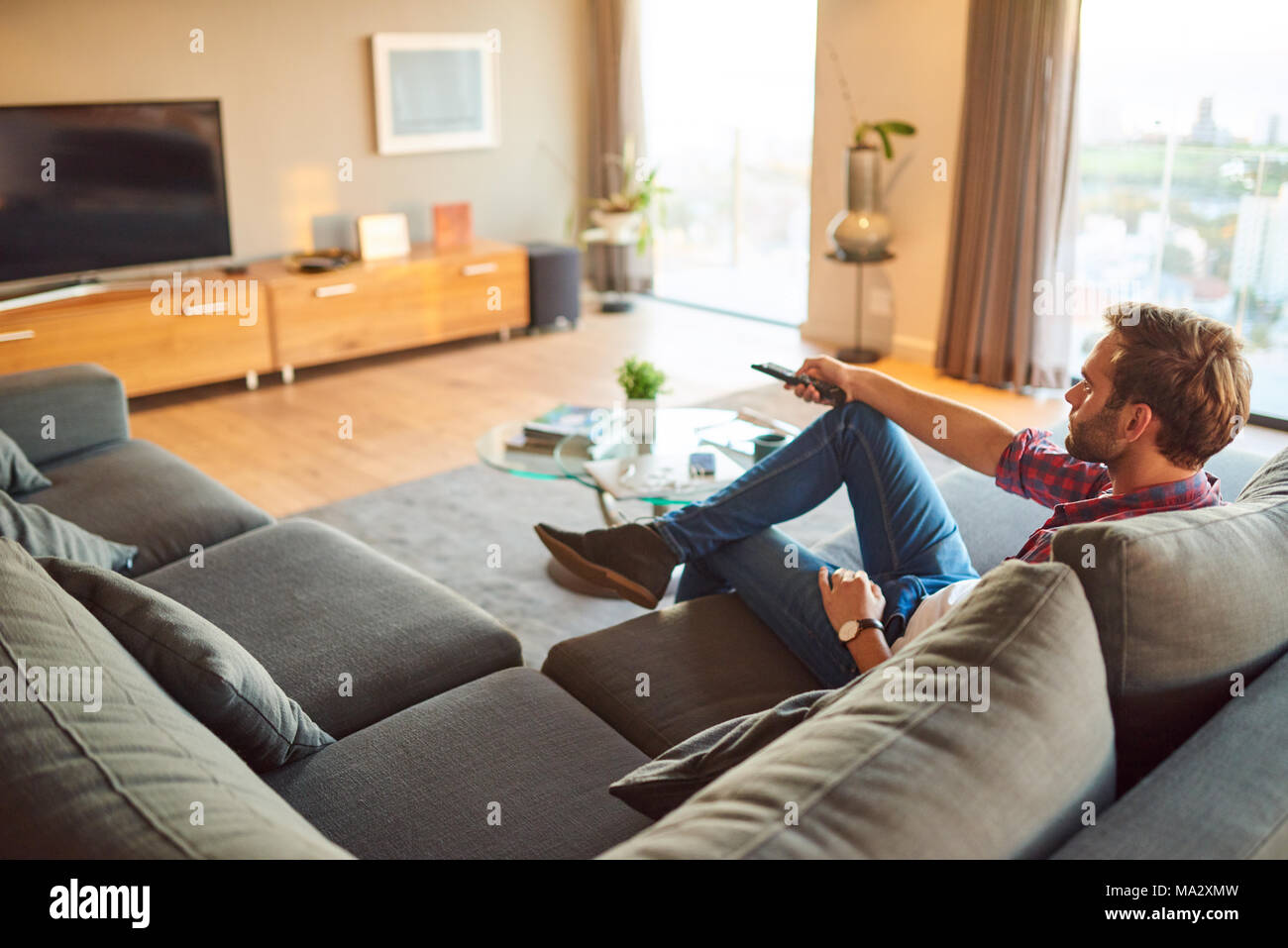 Young caucasian man sitting in his amazing modern apartment's living room, watching TV and changing the channel with the remote he is holding in his h Stock Photo