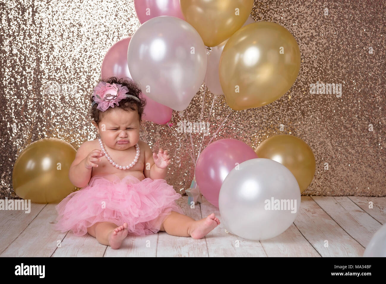 An unhappy, one year old, baby girl sitting with a bunch of balloons. She is wearing a pink tutu and a string of pearls. Stock Photo