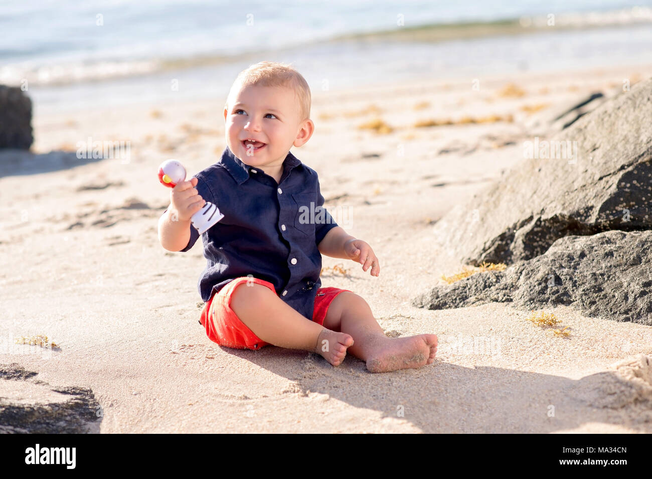 A one year old baby boy sitting on a beach and playing with a shaker toy. Stock Photo