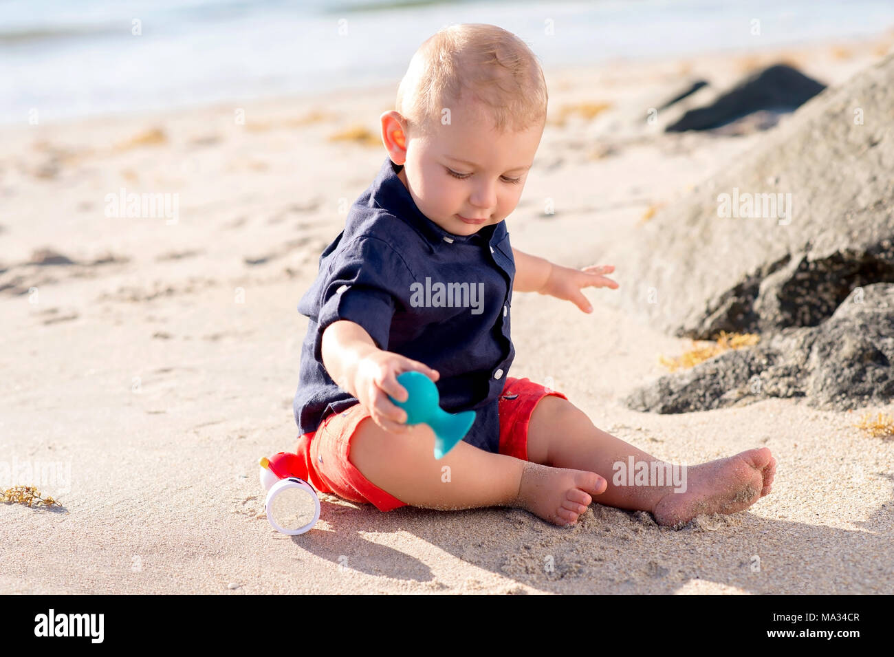 A one year old baby boy sitting on a beach and playing with a toy. Stock Photo