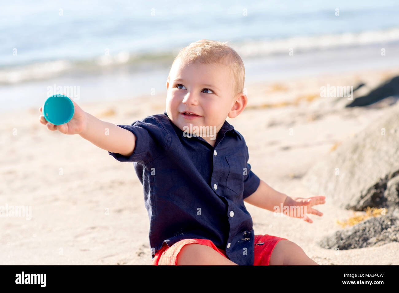 A one year old baby boy sitting on a beach. He is smiling and handing his toy to someone off camera. Stock Photo
