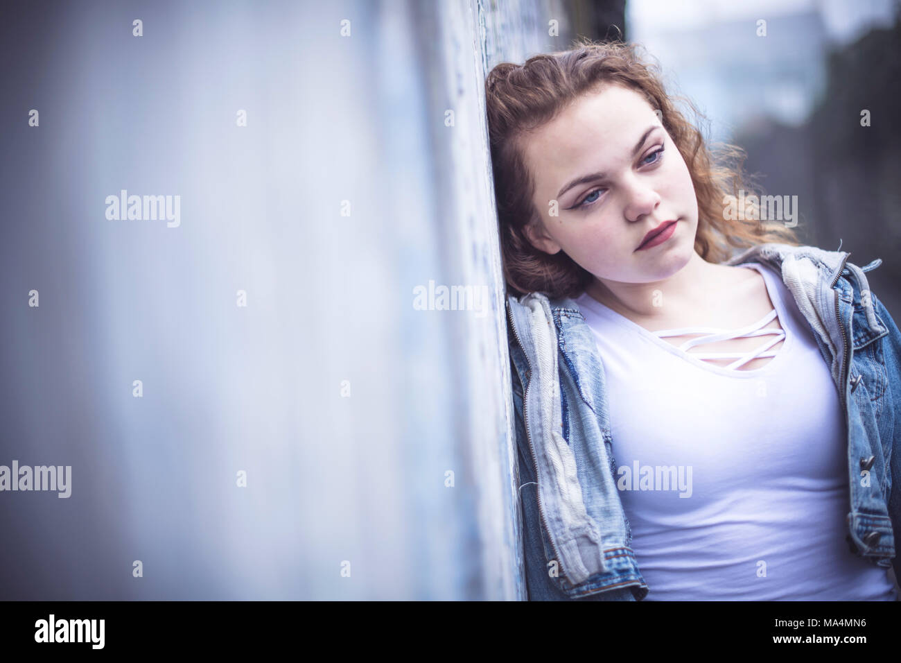 A moody attrractive young 13 thirteen year old teenage adolescent girl wearing a denim jacket, in an urban city back alley , UK Stock Photo
