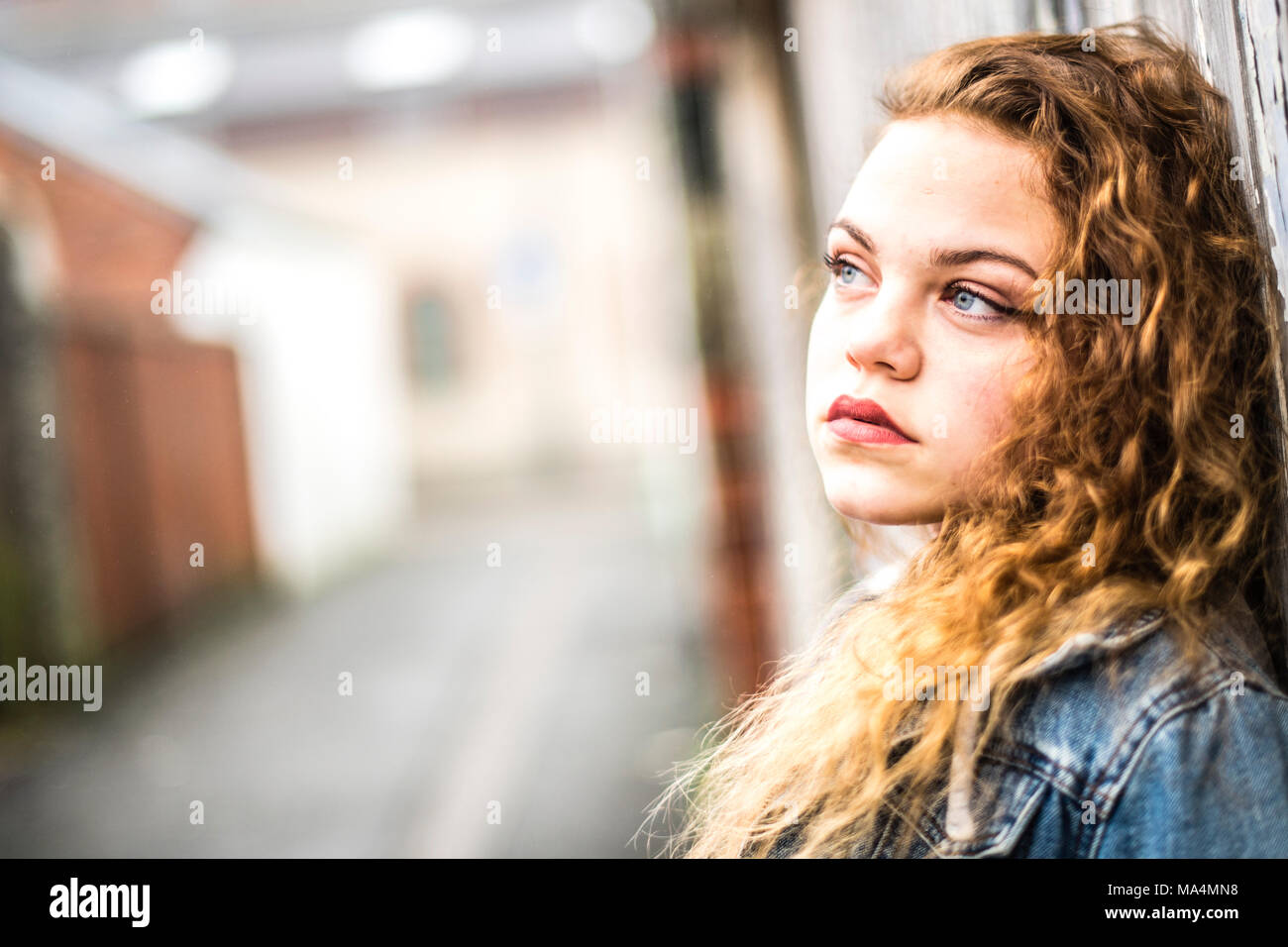 A moody attrractive young 13 thirteen year old teenage adolescent girl wearing a denim jacket, in an urban city back alley , UK Stock Photo