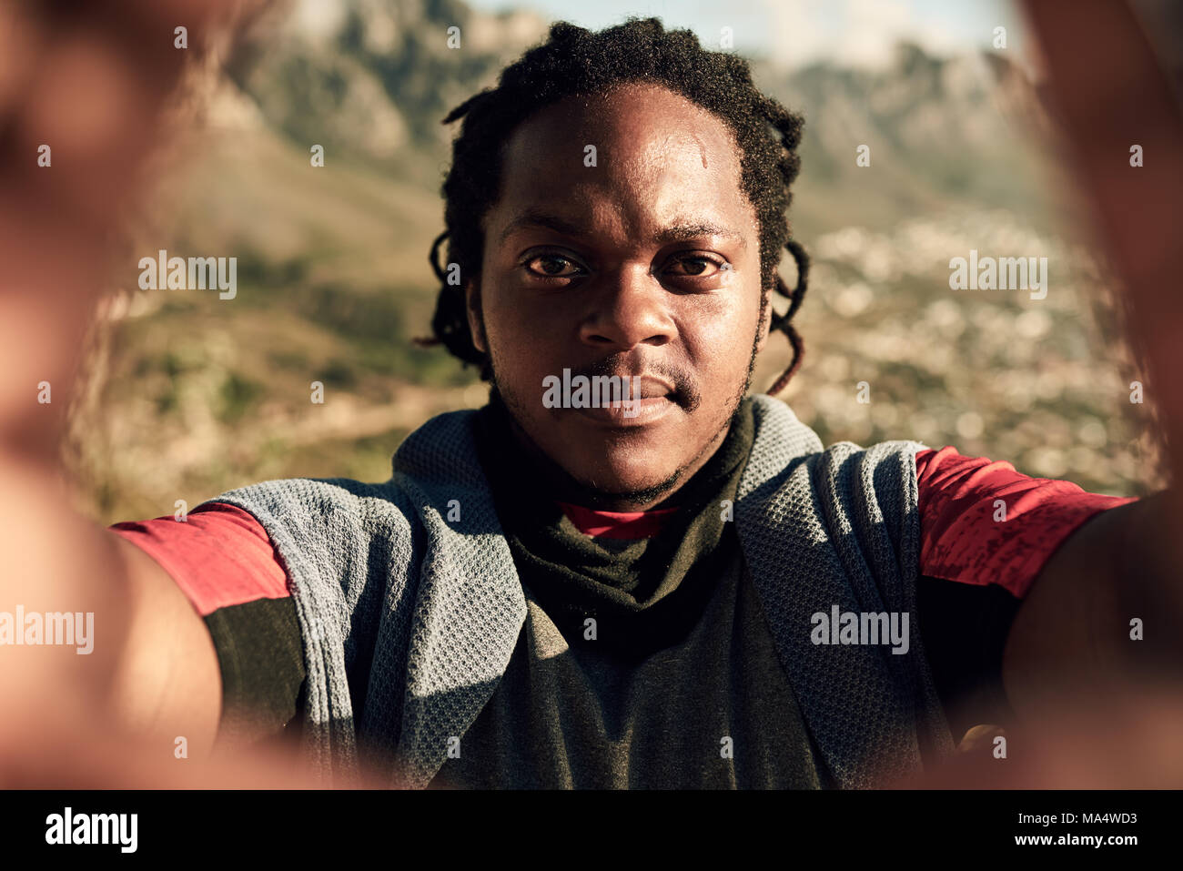 Attractive young african guy taking a selfie while on an extreme hike, scaling the side of a rather large mountain as the sun sets on a summer evening Stock Photo