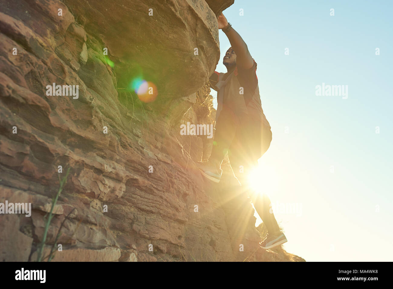 African guy climbing a near vertical rock face with clear blue skies and flare from the sun shining through from behind the climber. Stock Photo