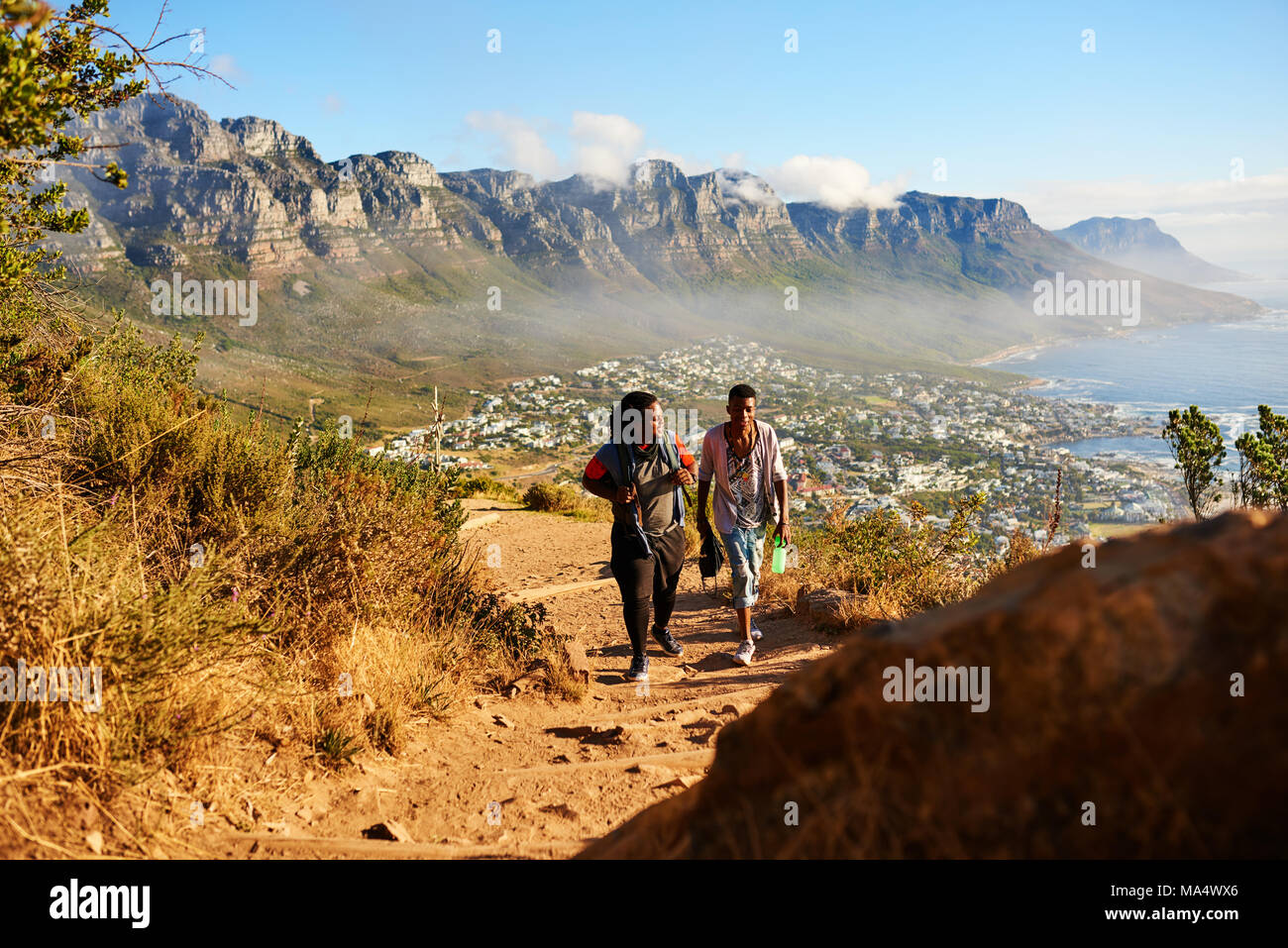 Two black male friends hiking a mountainous trail with a stunning scene of mountains, ocean and city scape behind them, as they ascend the mountain, c Stock Photo