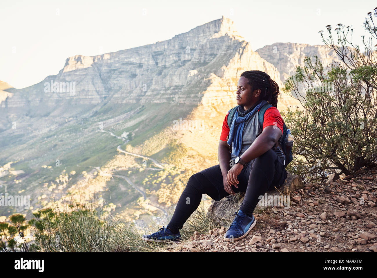 African hiker taking a break sitting next to the hiking trail on a mountainous trail to take in the amazing views of all the mountains around him that Stock Photo