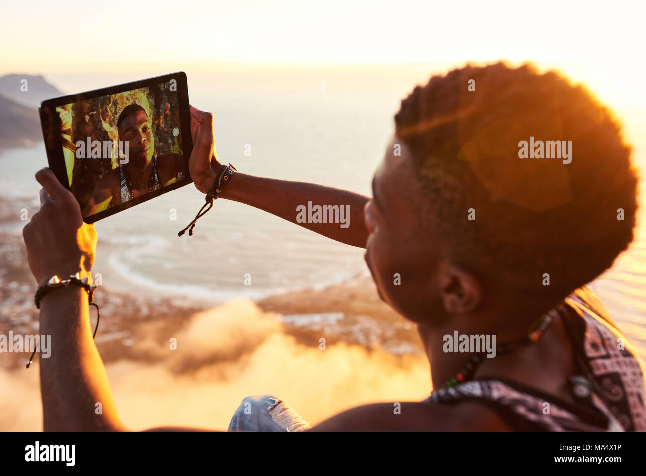 Young stylish black teenager busy using a tablet to take a selfie of himself on the top of a mountain trail while hiking on mountainous terrain on a w Stock Photo